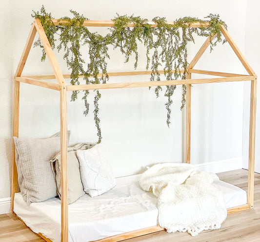 Wooden children's bed with a canopy and decorative plants, set against a white wall.