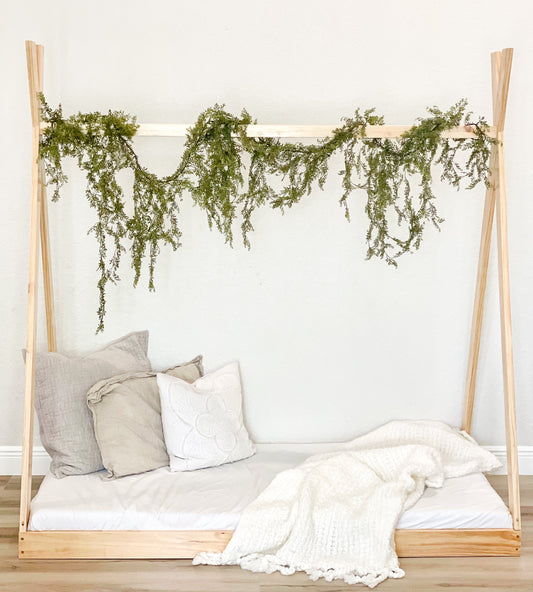Wooden bed frame with greenery, pillows, and a blanket against a white wall.